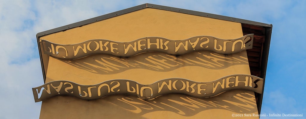 Palazzo delle ombre: una serie di onde metalliche bucate riflettono sul muro la parola "Più" in diverse lingue
